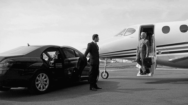 Businessman exits a private jet while a chauffeur holds the car door open on an airport runway.